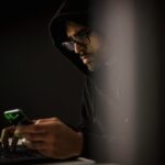 Online stalker in dark room using phone and laptop, representing digital harassment and cyberstalking threat